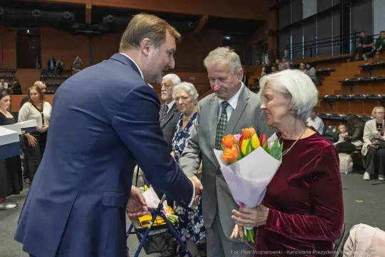 Mężczyzna w garniturze wita się z parą starszych osób, kobieta trzyma bukiet tulipanów.