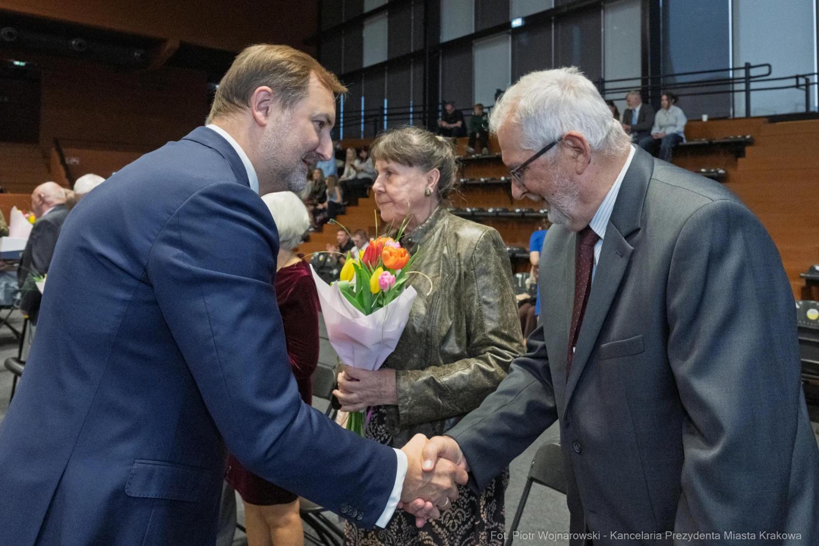 Uścisk dłoni między mężczyzną a starszym panem, w tle kobieta z bukietem kwiatów.  Autor: Piotr Wojnarowski