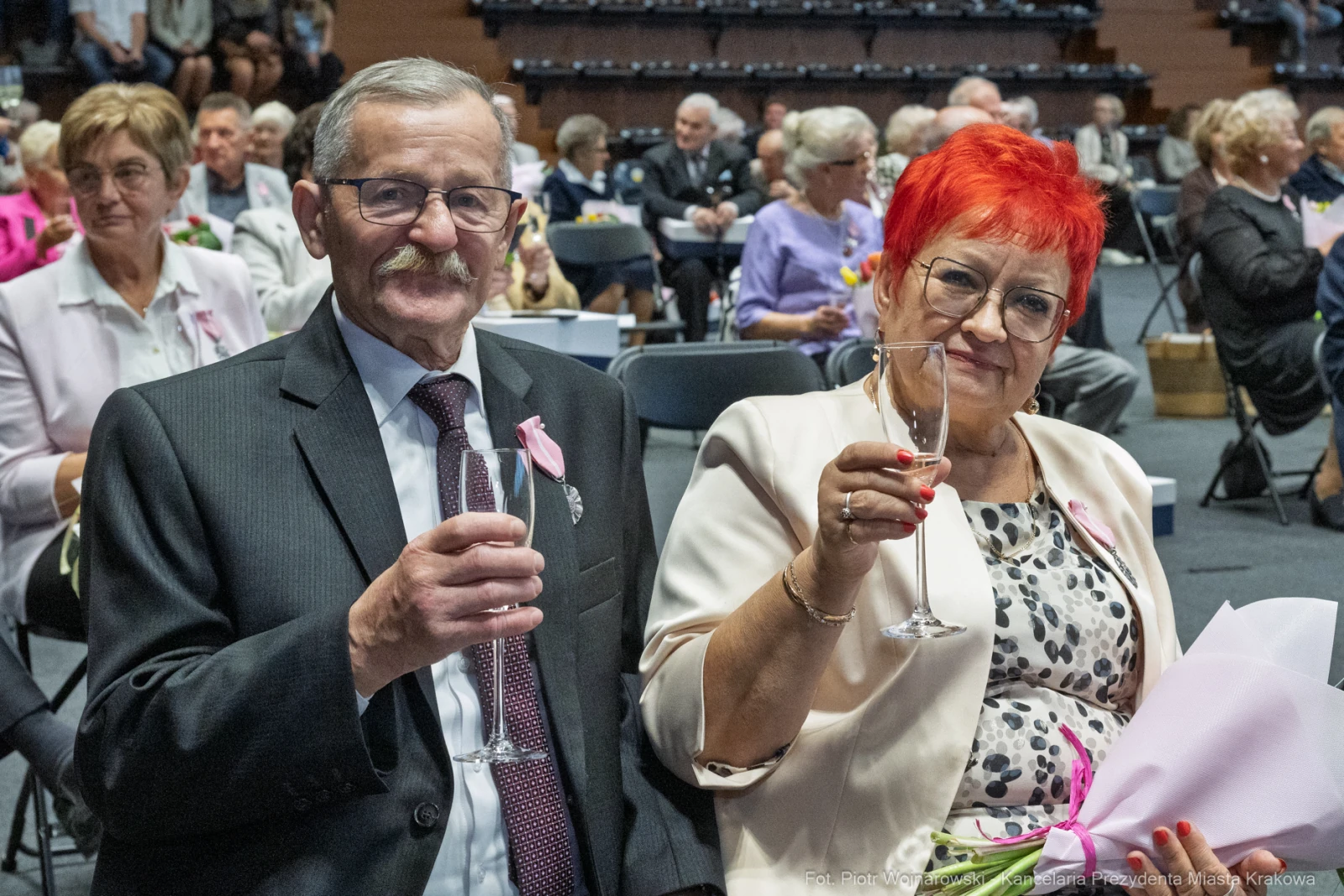 Starsza para w eleganckich strojach trzyma kieliszki, uśmiechając się do kamery.  Autor: Piotr Wojnarowski