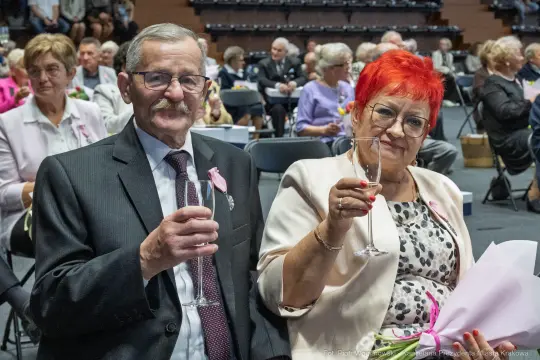 Starsza para w eleganckich strojach trzyma kieliszki, uśmiechając się do kamery.