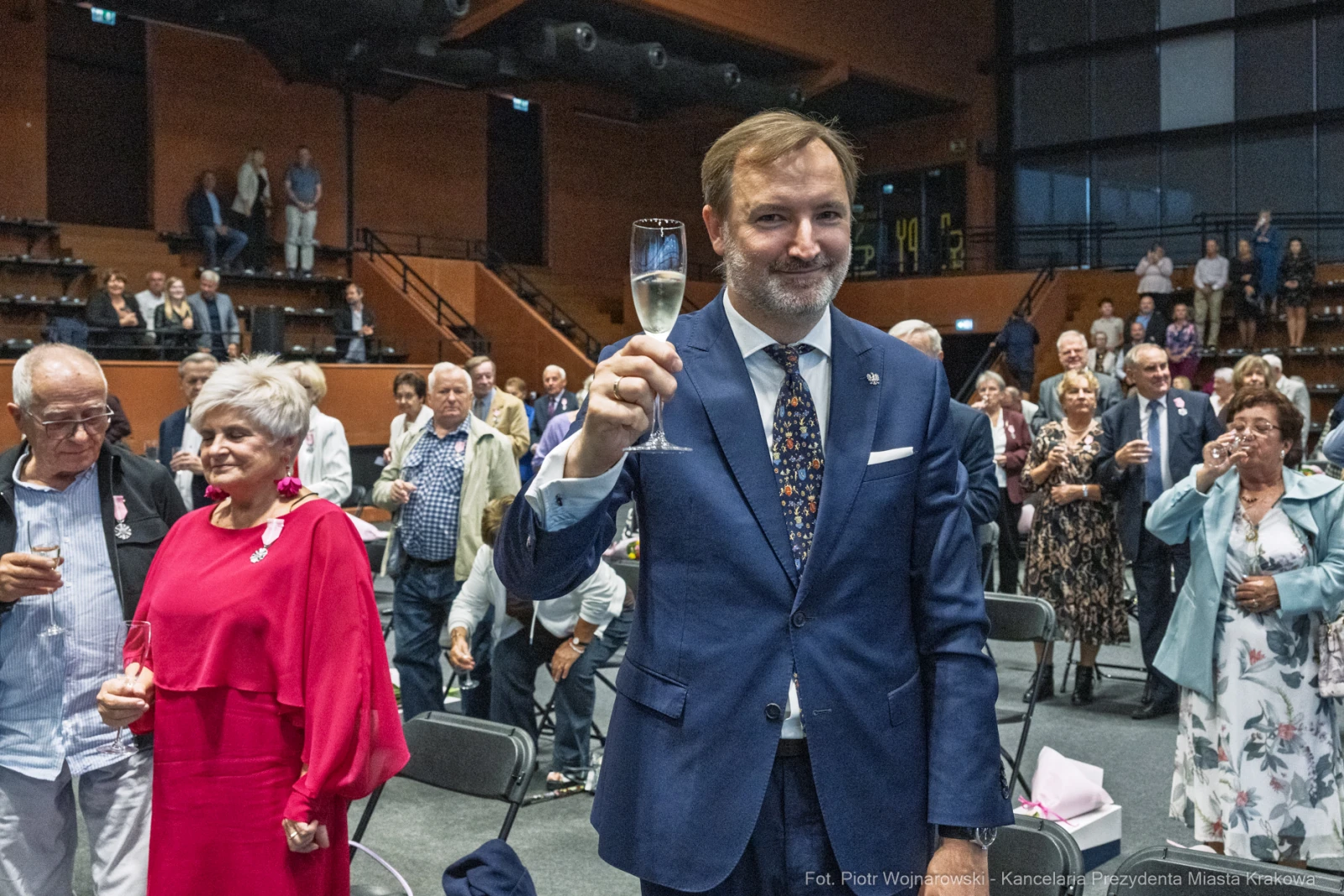 Mężczyzna w garniturze wznosi kieliszek szampana w towarzystwie gości na uroczystości.  Autor: Piotr Wojnarowski