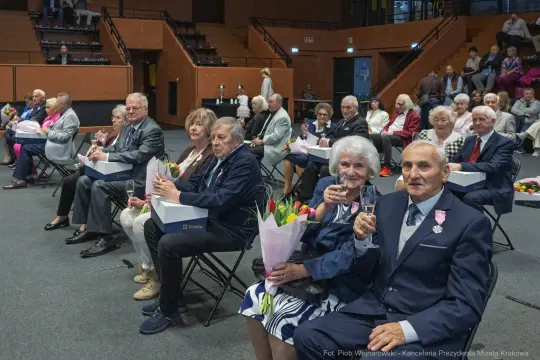 Uroczystość z okazji wręczenia nagród, uczestnicy siedzą z bukietami kwiatów i szklankami.