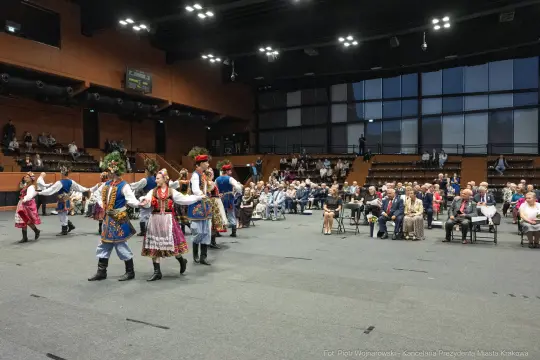 Grupa tancerzy w tradycyjnych strojach ludowych tańczy na scenie, publiczność obserwuje.