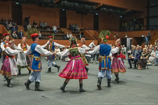 Tancerze w tradycyjnych strojach ludowych tańczą w kręgu na scenie.