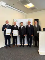 Czterech mężczyzn w garniturach trzymających dokumenty, stojących przed banerem z logo organizacji.