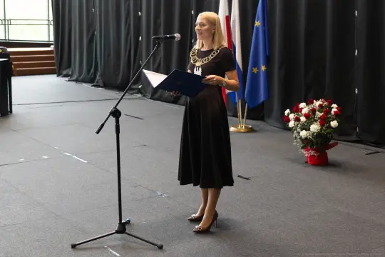 Kobieta w czarnej sukience przemawia do mikrofonu, obok flagi i bukietu kwiatów.