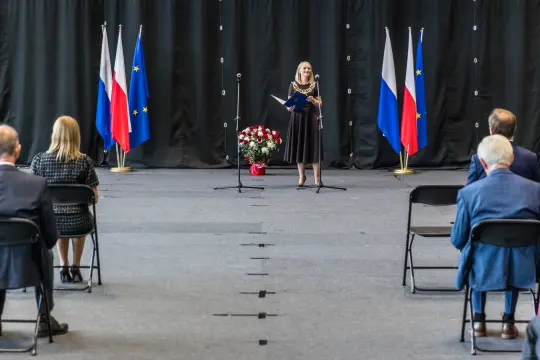 Kobieta w eleganckiej sukience przemawia na tle flag Polski i Unii Europejskiej.