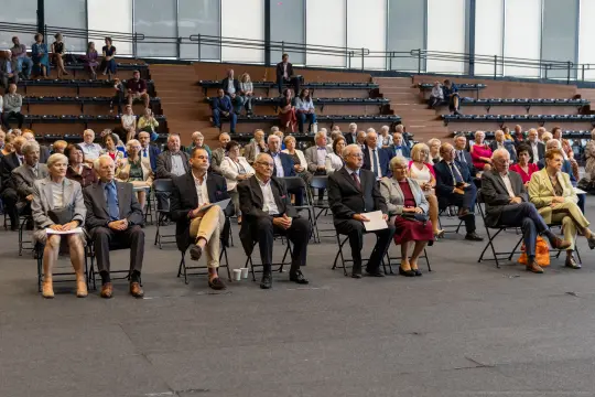 Grupa ludzi siedzących na krzesłach w dużej sali, uczestniczących w wydarzeniu.