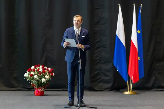 Mężczyzna w garniturze przemawia przy mikrofonie, obok flagi Polski i Unii Europejskiej oraz kwiaty.