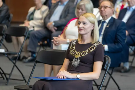 Kobieta w eleganckiej sukience z łańcuchem na szyi siedzi na krześle wśród publiczności.