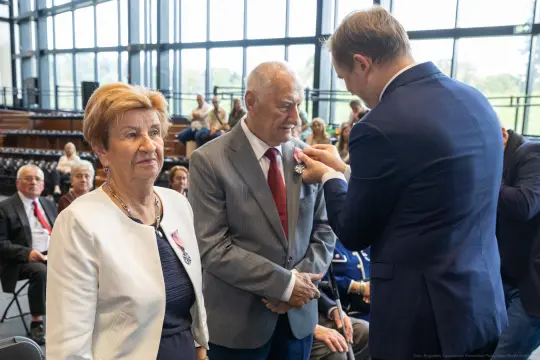 Uroczystość wręczenia odznaczeń, dwie osoby w eleganckich strojach, mężczyzna odbiera medal.