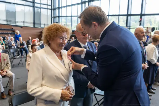 Wręczenie odznaczenia kobiecie w białym garniturze podczas uroczystości.