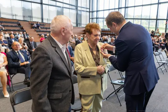Uroczystość wręczenia odznaczeń, starsza kobieta odbiera medal od mężczyzny. W tle zgromadzenie gości.