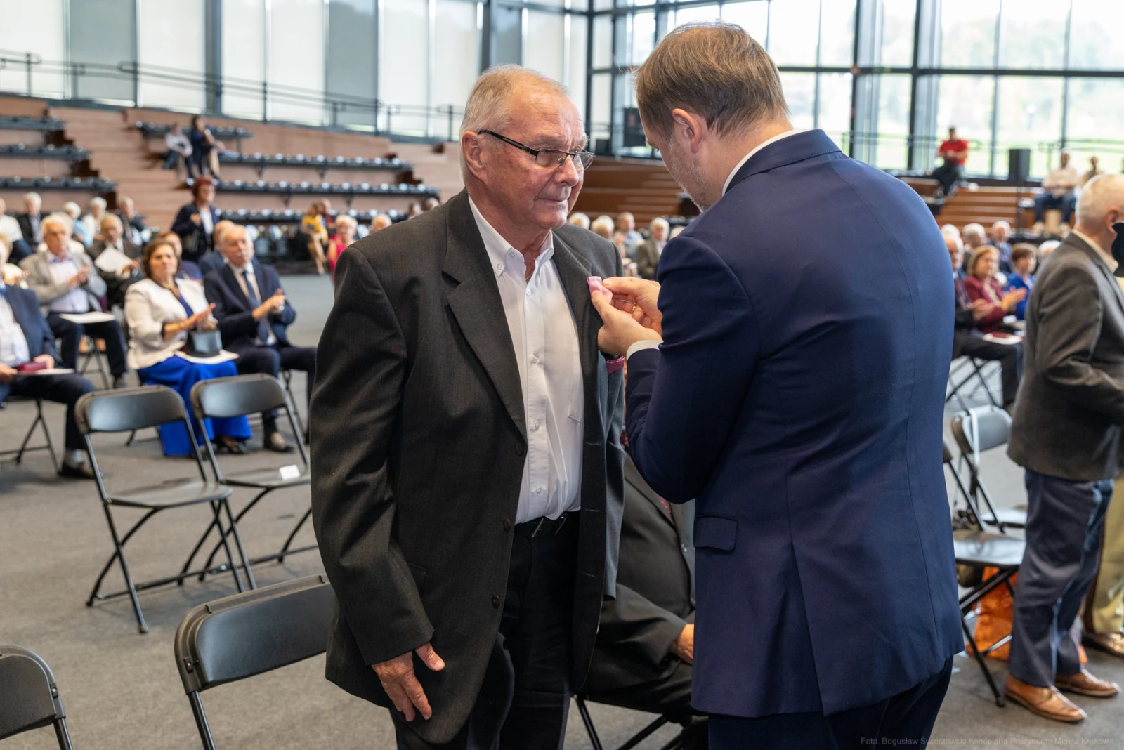 Mężczyzna odbiera odznaczenie podczas uroczystości w sali z siedzącymi gośćmi.  Autor: Bogusław Świerzowski