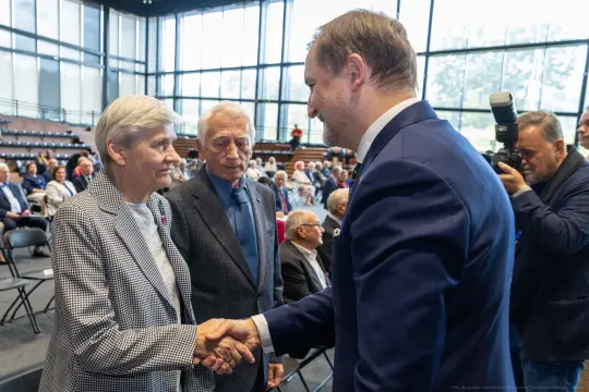 Spotkanie dwóch osób z mężczyzną w garniturze, uściski dłoni, w tle publiczność.