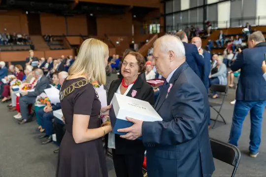 Wręczenie nagrody przez kobietę starszym osobom w sali wypełnionej gośćmi.
