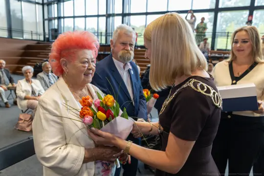 Kobieta z różowymi włosami otrzymuje bukiet kwiatów od młodszej kobiety. W tle goście w sali.