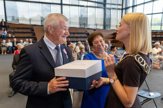 Uroczystość wręczenia nagrody, mężczyzna z uśmiechem trzyma pudełko, obok kobieta w okularach.