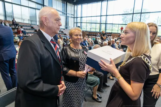 Wręczenie nagrody przez kobietę starszym mężczyźnie i kobiecie w sali wypełnionej ludźmi.