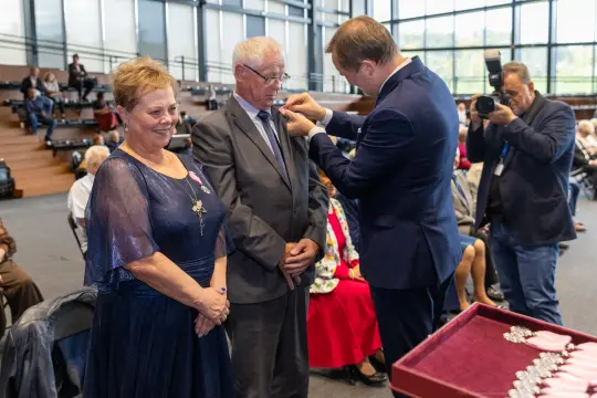 Wręczenie odznaczenia mężczyźnie w towarzystwie uśmiechniętej kobiety.