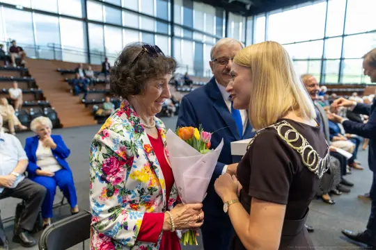 Kobieta z bukietem kwiatów rozmawia z inną kobietą w eleganckim otoczeniu.