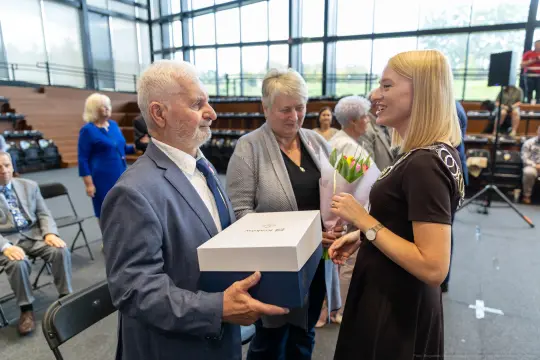 Wręczenie nagrody przez kobietę mężczyźnie, w tle widoczni inni uczestnicy wydarzenia.