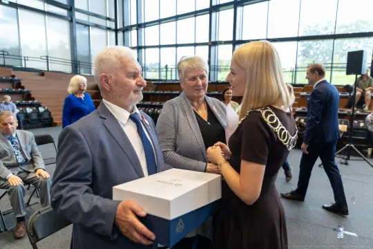 Mężczyzna wręcza kobietę pudełko, obok stoi inna kobieta, w tle widoczna sala z publicznością.