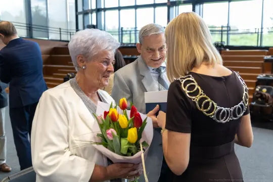 Starsza para z bukietem tulipanów rozmawia z kobietą w eleganckiej sukience.