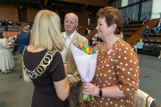 Para z bukietem tulipanów rozmawia z osobą w łańcuchu. W tle widoczna publiczność.