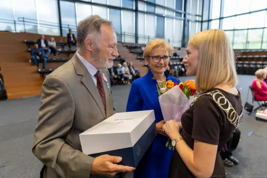 Mężczyzna wręcza kobietom kwiaty i prezent w sali pełnej widowni.