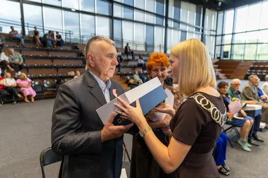 Wręczenie odznaczenia mężczyźnie przez kobietę w eleganckim stroju, w tle siedzący tłum.