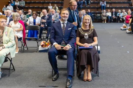 Uroczystość z uczestnikami siedzącymi na krzesłach, w tle widoczni goście.