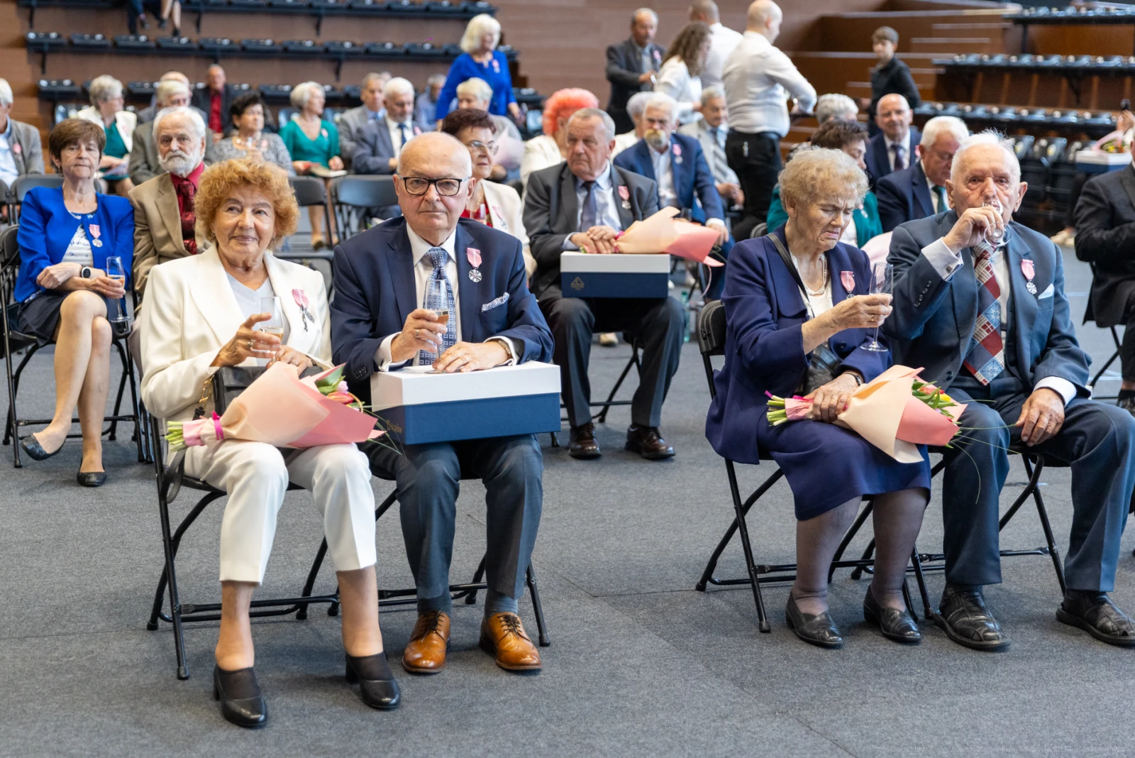 Uczestnicy uroczystości siedzą na krzesłach, trzymając kwiaty i napoje.  Autor: Bogusław Świerzowski