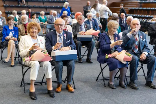 Uczestnicy uroczystości siedzą na krzesłach, trzymając kwiaty i napoje.