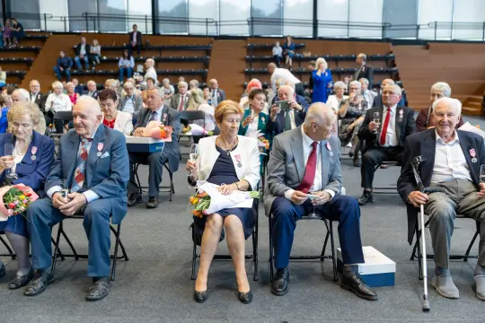 Grupa starszych osób siedzących w sali, niektórzy z medalami, podczas uroczystości.