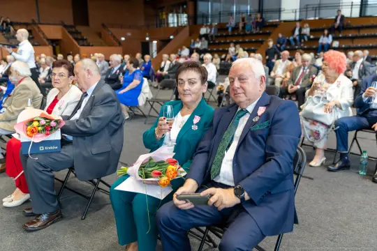Uczestnicy uroczystości siedzący na sali, trzymający kwiaty i medale.