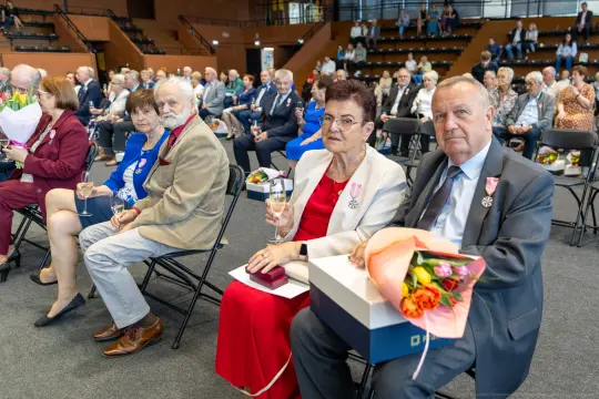 Uczestnicy uroczystości siedzący na widowni, trzymający kwiaty i kieliszki.