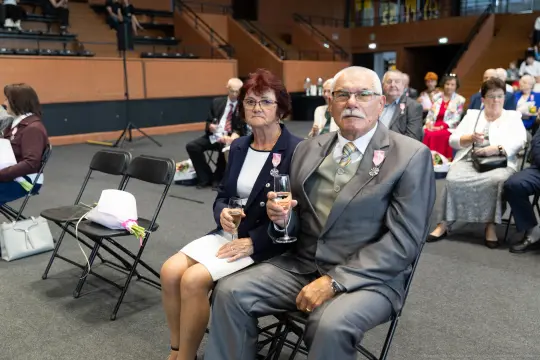 Starsza para siedząca na ceremonii, trzymająca kieliszki. W tle inni goście w formalnych strojach.