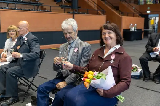 Uczestnicy ceremonii z medalami, trzymający kieliszki i bukiety kwiatów.