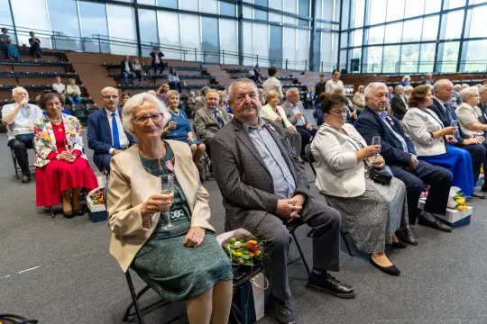 Grupa osób w eleganckich strojach siedzi na widowni, trzymając kieliszki.