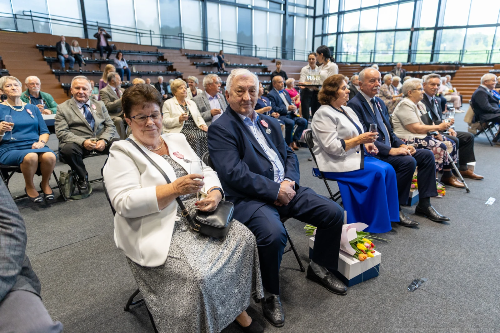 Grupa osób siedzących na widowni, ubranych elegancko, z uśmiechami i kieliszkami w rękach.  Autor: Bogusław Świerzowski