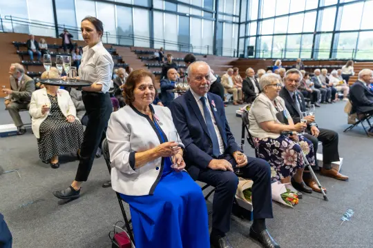 Uczestnicy uroczystości siedzą na widowni, trzymając kieliszki z napojami.