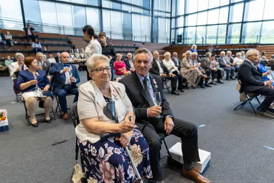 Uczestnicy uroczystości siedzący na krzesłach, trzymający kieliszki.