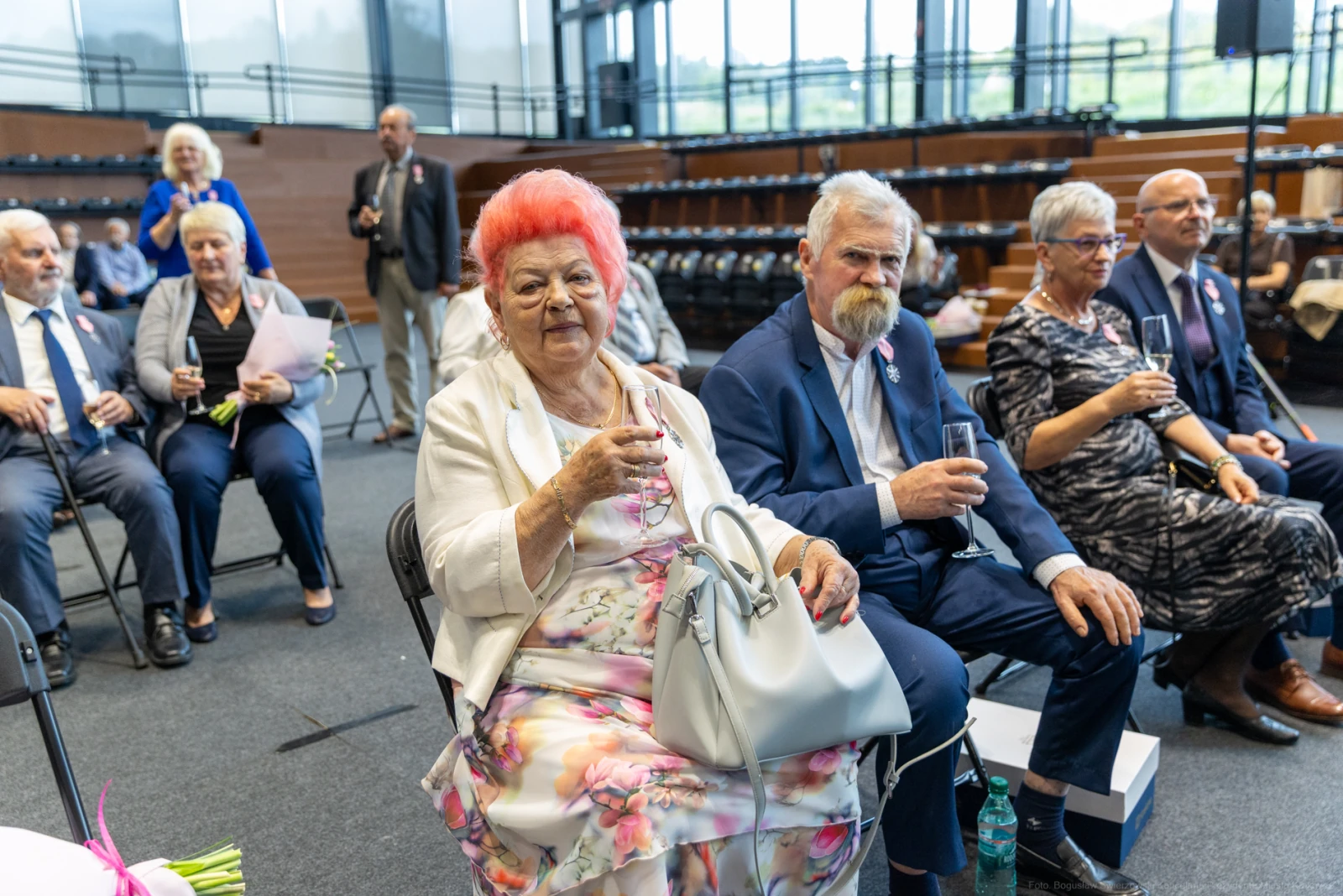 Grupa osób w eleganckich strojach, część z kieliszkami, siedzi w sali.  Autor: Bogusław Świerzowski