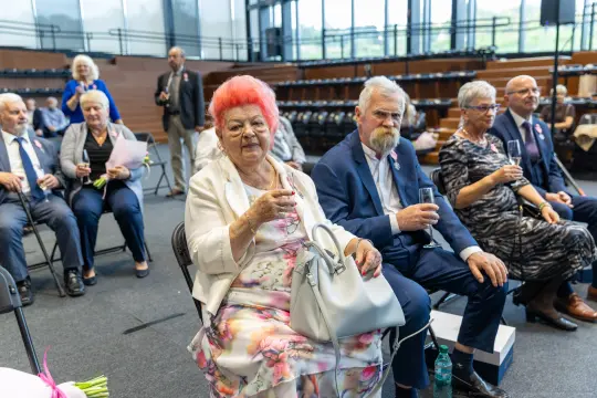 Grupa osób w eleganckich strojach, część z kieliszkami, siedzi w sali.