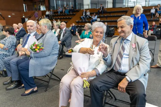 Uczestnicy uroczystości z medalami, wznoszący toast i trzymający kwiaty.