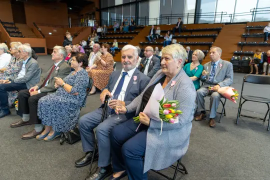 Grupa osób siedzących na widowni, niektórzy trzymają kwiaty i kieliszki.
