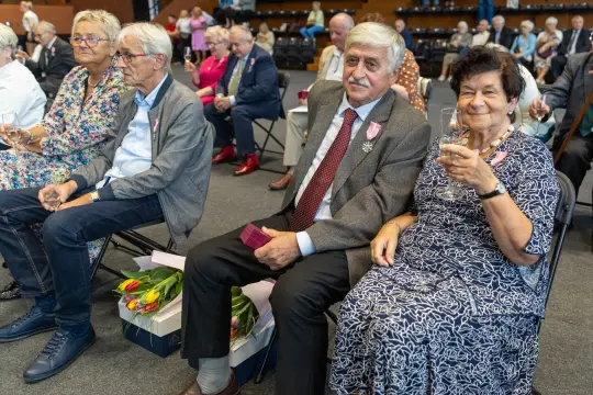 Uczestnicy uroczystości siedzący na krzesłach, trzymający kieliszki i kwiaty.