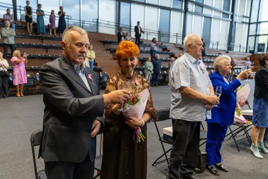Uczestnicy uroczystości w eleganckich strojach, wznoszący kieliszki i trzymający kwiaty.
