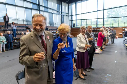 Uczestnicy uroczystości z kieliszkami w rękach, uśmiechnięci i w eleganckich strojach.